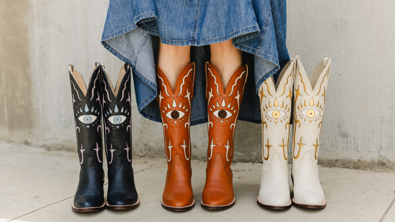 Four decorative cowboy boots displayed side by side on a concrete surface, with a person's legs and blue skirt visible above them. The boots are navy, brown, and white, each featuring a large stylized evil eye motif and western embroidery.
